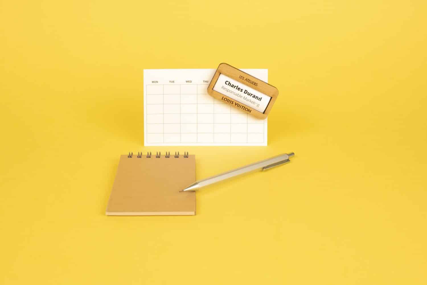 Un calendrier avec un stylo et un bloc-notes sur fond jaune avec un Badge rectangle en bois et français.