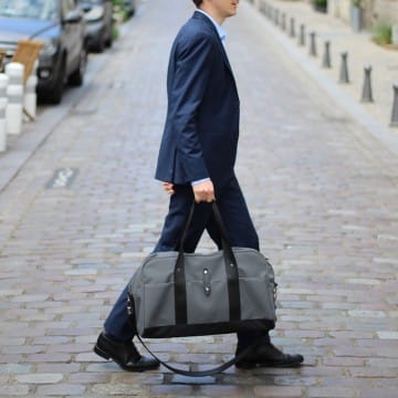 Un homme en costume marchant dans une rue pavée portant un sac polochon, présentant un Sac week-end en toile issu de l'upcycling et fabriqué en France.
